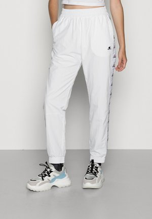Persoon draagt witte trainingsbroek met elastische taille, logoband aan de zijkant en stevige witte, grijze en blauwe sneakers op een grijze vloer.