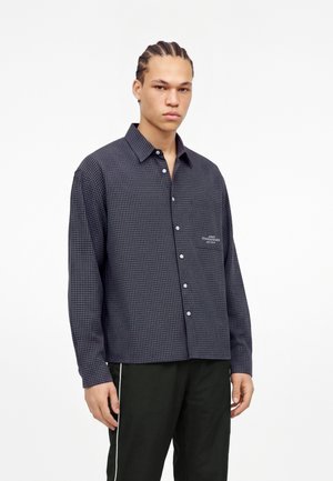 Jeune homme avec une coiffure en cornrows porte une chemise à carreaux sombre avec boutons et un pantalon noir avec une bande blanche sur un fond blanc uni.