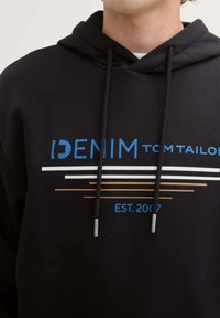 Sudadera negra con un gráfico de texto en azul y blanco, líneas horizontales en naranja y blanco, y cordones con puntas metálicas.