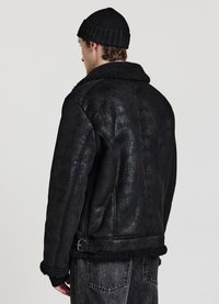 Giacca in shearling nero con ampio colletto, tessuto lavorato e dettaglio con fibbia in vita. Indossata con berretto in maglia nero e jeans grigi.