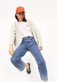 Gorra de béisbol naranja, blazer de lino beige, camiseta blanca con logo, vaqueros de pierna ancha azules, zapatillas negras, pose sentada contra un fondo blanco.