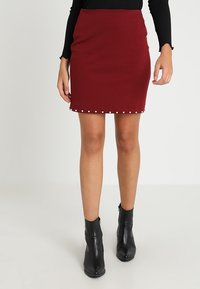 Burgundy kokerrok met een textuurafwerking en witte decoratieve studs langs de zoom; gedragen met zwarte enkellaarzen.
