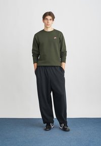 Jovem em pé com as mãos nos bolsos, vestindo um fato de treino verde-azeitona da Nike, calças largas pretas e sapatilhas pretas em um chão azul.