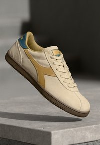 Tênis de couro bege com uma faixa lateral amarela, detalhe azul no calcanhar, bico redondo e sola de borracha texturizada, apresentando um design clássico com atacadores.