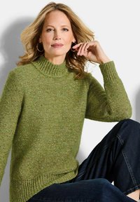Grüner Strickpullover mit hochgeschlossenem Ausschnitt und gerippten Akzenten. Hat ein strukturiertes Muster. Wird mit dunkelblauen Jeans getragen.
