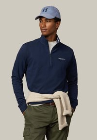 Felpa con zip blu navy con colletto interno bianco, maglione beige annodato in vita, pantaloni cargo verdi e cappellino azzurro chiaro con logo "H".
