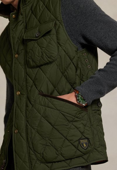 Persona que lleva un chaleco acolchado verde con la mano en el bolsillo y un reloj verde, combinado con una camisa de manga larga gris oscuro.