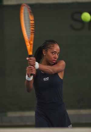 Tennisspeler in een blauwe mouwloze top en rok, met een oranje racket, klaar om een gele tennisbal te slaan. Achtergrond van een binnenbaan.