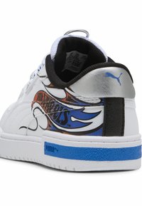 Zapatilla blanca con piel blanca texturizada, acentos negros y un talón plateado brillante. Presenta un diseño gráfico colorido y un detalle en la entresuela de color azul.