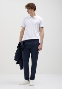 Giovane che indossa una polo bianca, pantaloni blu scuro e sneaker bianche tiene una giacca blu scuro, guardando verso il basso a sinistra.