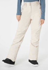 Pantaloni da sci beige chiaro con vita alta, due tasche frontali, chiusura a tre bottoni e gamba affusolata; abbinati a stivali da neve neri.