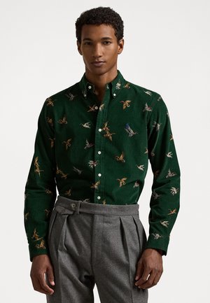 Groene corduroy shirt met geborduurde eenden, met knopen en lange mouwen. Gecombineerd met grijze geplooide broeken.