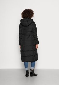 Abrigo negro acolchado con capucha, con secciones acolchadas y una silueta larga. Llevado sobre unos vaqueros azules remangados y botas negras de tobillo.