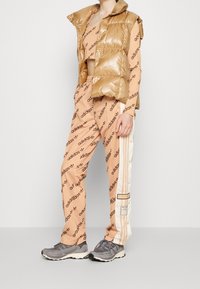 Osoba oblečená v béžovém teplákovém soupraven Adidas s logo potiskem, bílými bočními pruhy, péřovou vestou a šedými běžeckými teniskami, stojící na jednobarevném pozadí.