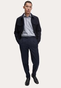Giacca in misto lana navy sopra una camicia a quadri azzurri chiari, abbinata a pantaloni navy su misura e scarpe eleganti nere lucidate.