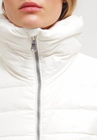 Bílá vyp padded bunda s vysokým límcem, vybavená předním zipem a hladkým lesklým povrchem. Stříbrné kování zdobí design.