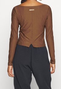 Žena viděná ze zadu v přiléhavém hnědém halenkovém tričku Nike Air s dlouhým rukávem a volných černých fleecových kalhotách na jednoduchém pozadí.