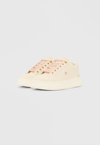 Bež superge z roza vezalkami, gladkim sintetičnim zgornjim delom, zaobljenim prstom in debelim belim podplatom. Na strani so diskretno vidni logotipi.