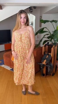 Jeune femme aux longs cheveux debout à l’intérieur, portant une robe fleurie sans manches et des ballerines grises à côté d’un canapé marron et d’un appareil d’exercice.
