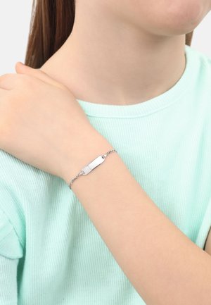 Pulsera de plata con un charm plano y rectangular, unida por una delicada cadena. Llevada en una muñeca, contra un suéter de punto verde menta.