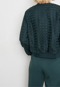 Person med mörkgrön byxor och en strukturerad mörkgrön jacka med geometriska mönster, sedd bakifrån mot en vit bakgrund.