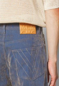 Modré džíny z denimu s texturovaným povrchem, vybavené velkou, embosovanou hnědou koženou štítkem a výrazným cik-cak vzorem na zadní kapse.