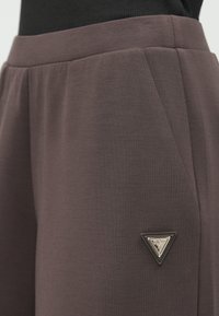 Nahaufnahme einer dunkelbraunen Hosentasche mit einem kleinen dreieckigen Guess-Logo-Patch an der Seite und einem schwarzen Bund.