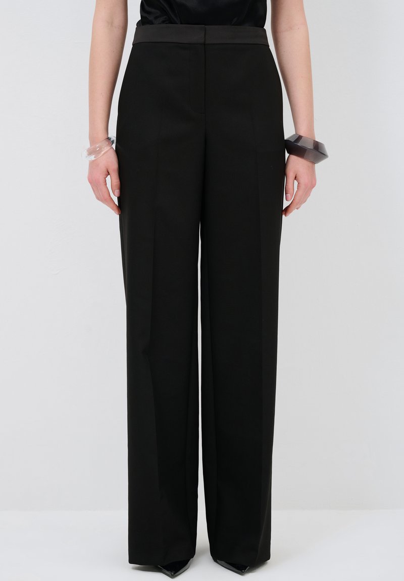 Pantalons larges noirs avec une taille haute, confectionnés en tissu lisse. Présentent des plis marqués et une silhouette ajustée.