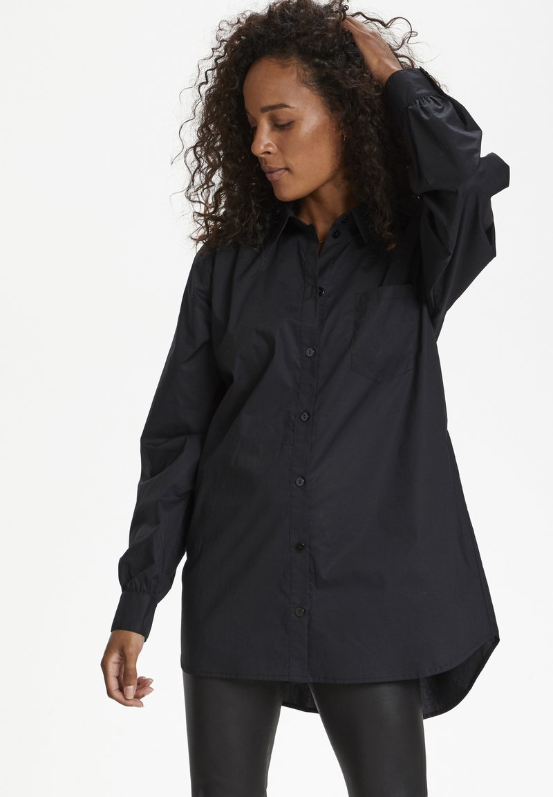 Black button-up shirt with a relaxed fit, long sleeves, and a chest pocket. Made from smooth fabric with a slight sheen and curved hemline.