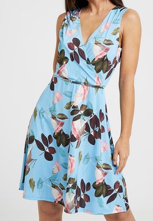 Bezrekawowa jasnoniebieska sukienka w różowo-brązowy wzór kwiatowy, z dekoltem w serek i dopasowaną talią na stojącej kobiecie.