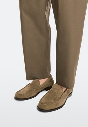 Braune Wildleder-Slipper, getragen mit passenden braunen Hosen, zeigen Nahtdetails und schwarze Sohlen auf weißem Hintergrund.