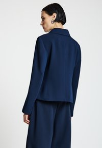 Blazer bleu marine en tissu texturé, avec un col à revers cranté et une coupe ajustée. Le dos présente des lignes épurées et des manches longues.