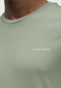 Κοντινό πλάνο ατόμου που φοράει γαλάζιο-πράσινο μπλουζάκι Calvin Klein με λευκό λογότυπο στο στήθος, εμφανίζοντας τη γραμμή του λαιμού και το πάνω μέρος του σώματος.