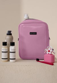 Roze hardshell reistas met geribbelde textuur, zwart logo, twee shampoo flesjes, een conditionerfles, een roze container met bloemenontwerp en een rode tandenborstel.