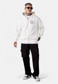 Hoodie zippé blanc avec un logo sur la poitrine, associé à un pantalon cargo noir avec de grandes poches et des baskets noir et blanc.