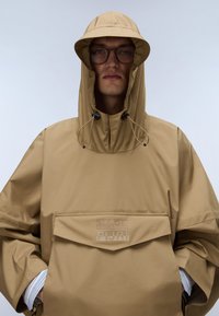 Jachetă impermeabilă de culoare maro deschis, cu glugă, șnururi și buzunar frontal; are un logo de marcă în partea de jos.