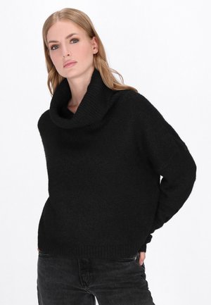 Donna con capelli biondi che indossa un maglione nero a collo alto e jeans scuri, in piedi con le mani nelle tasche su uno sfondo bianco.