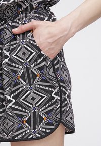 Tela negra con un patrón que presenta diseños geométricos en blanco, azul y naranja. Se puede ver un bolsillo lateral con una mano parcialmente insertada.