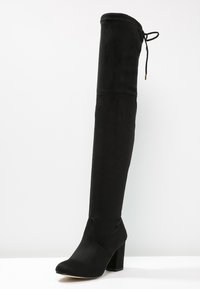 Bota negra sobre la rodilla hecha de tela suave, con un tacón grueso y un detalle de cordones en la parte superior, con una textura suave en toda su superficie.