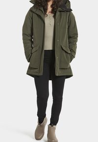 Parka olive verte avec capuche, deux poches avant et fermeture éclair, portée sur un haut beige et un pantalon noir, associée à des bottes beiges.