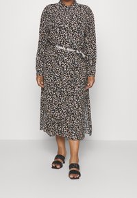Vestido midi negro con estampado floral, mangas largas, cuello con botón, cinturilla elástica y un cinturón blanco con logo. Sandalias de tiras negras completan el look.
