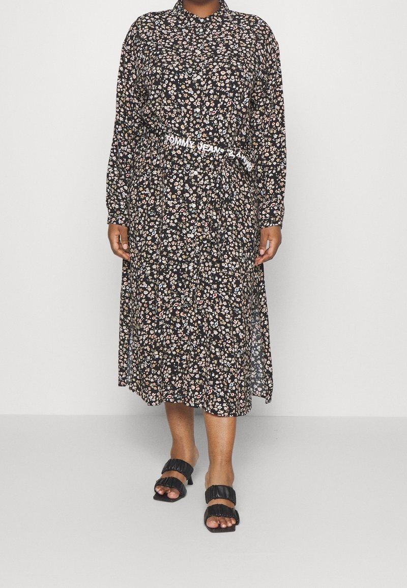 Vestido midi negro con estampado floral, mangas largas, cuello con botón, cinturilla elástica y un cinturón blanco con logo. Sandalias de tiras negras completan el look.