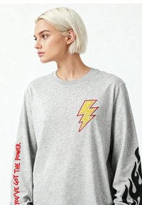 Junge Person mit kurzen blonden Haaren, die ein graues Sweatshirt mit gelbem Blitz, schwarzer Flammengrafik und rotem Text "Du hast die Kraft" trägt.