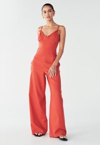 Žena v oranžovo-červenom bezrukávovom overal s širokými nohavicami a sandálmi, stojí pred jednoduchým bielym pozadím.