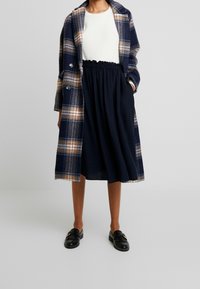 Rutet frakke i marineblå, beige og hvid; cremefarvet sweater; sort nederdel med elastisk talje; sorte loafers med dekorativ hardware; afslappet outfit.
