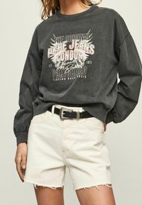 Sudadera gris oscuro con un diseño impreso que incluye texto y gráficos, combinada con pantalones cortos desgastados de un beige claro y un cinturón negro.