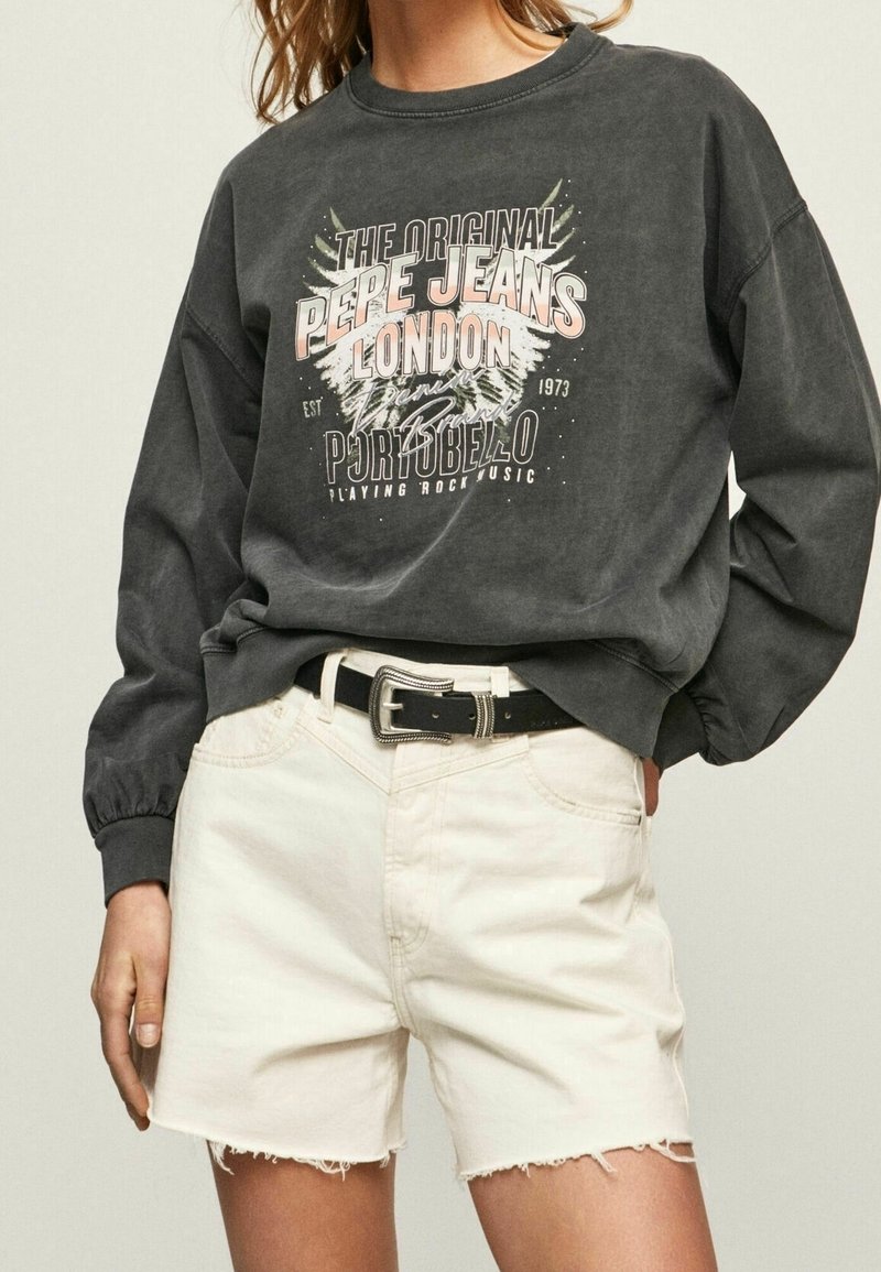 Sudadera gris oscuro con un diseño impreso que incluye texto y gráficos, combinada con pantalones cortos desgastados de un beige claro y un cinturón negro.