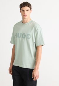 Mlad moški v svetlo zeleni majici s velikimi črkami "HUGO", v kombinaciji s črnimi hlačami, stoji pred enobarvno belo ozadje.
