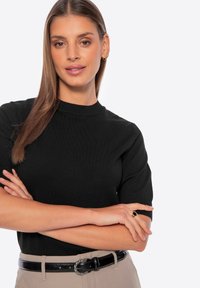 Černý pletený top s krátkými rukávy, s kulatým výstřihem. Model má na sobě béžové kalhoty a černý pás se stříbrnou sponou, ruce zkřížené.