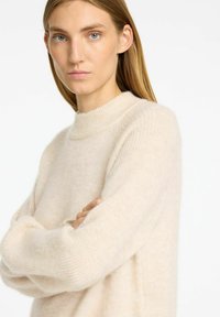 Beige stickad tröja med hög krage, sänkta axlar och mjuk textur, med en avslappnad passform och en avslappnad design.